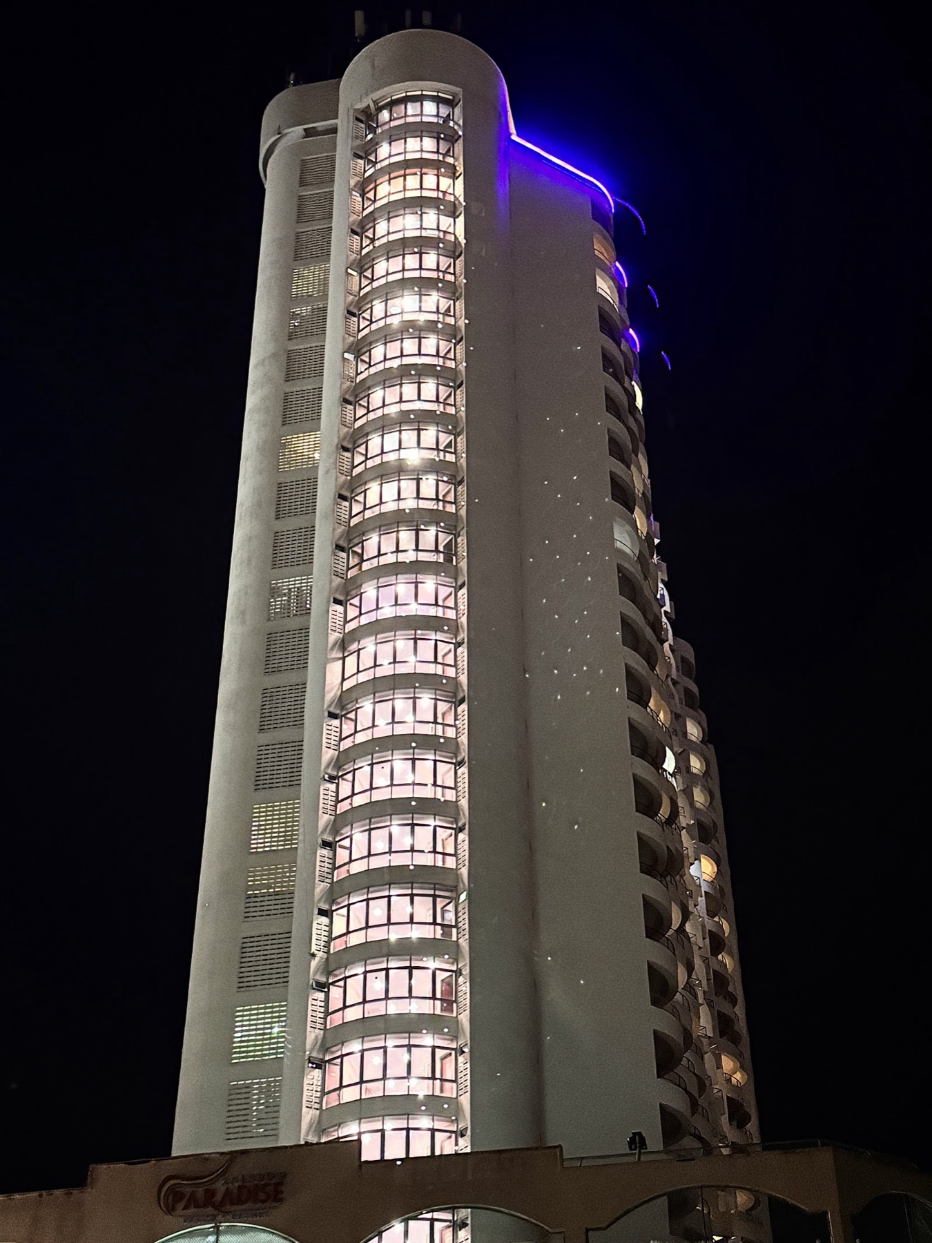 The curved residential tower of Rainbow Paradise lit pink and violet against the night sky