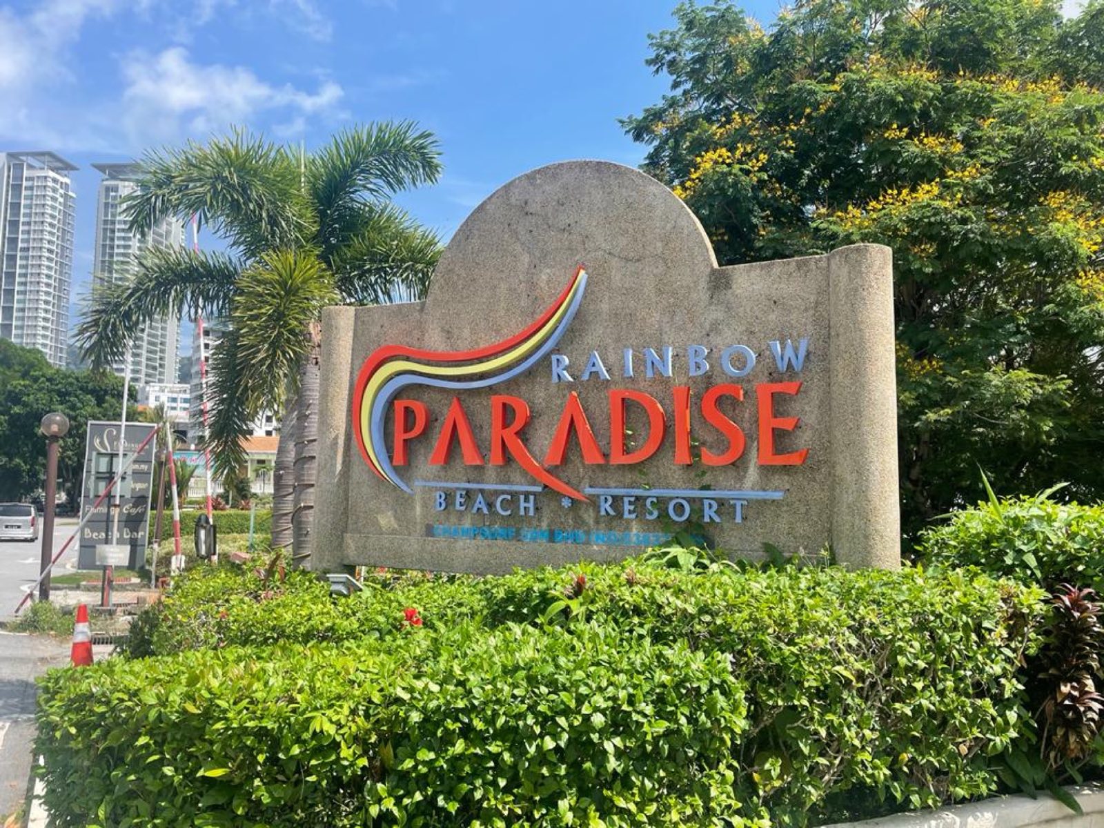 The Rainbow Paradise Beach Resort entrance sign, a rainbow wordmark set in stone among palms